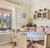 A cozy dining nook with a round table, wicker chairs, and a floral tablecloth. A window with pink curtains overlooks a grassy yard. Shelves and a wall clock decorate the beige wall.