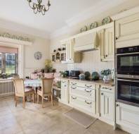 A cozy kitchen with cream cabinets, a wooden dining table, four chairs, built-in double ovens, and decorative plates on top. A window with a view of a garden lets in natural light.