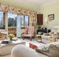 Elegant living room with large windows, floral curtains, armchairs, and decor. Sunlit view of outdoor greenery. Cozy and classic atmosphere.