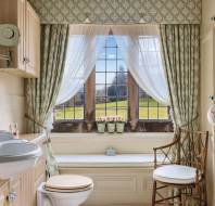 Traditional bathroom with a vintage sink and toilet, patterned wallpaper and curtains, wooden chair, and a large window overlooking a garden.