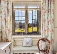 Interior view of a room with floral curtains framing a window. There's a cushioned bench below, a wooden chair, and a light-colored dresser. Through the window, grass and blue sky are visible.