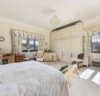 Spacious bedroom with a large bed, floral curtains, two chairs, a wooden table, and built-in wardrobes. Large windows allow natural light to fill the room. A chandelier hangs from the ceiling.