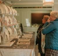 Two women look at jewelry displayed on white stands at an indoor market or exhibition, with other people browsing in the background.