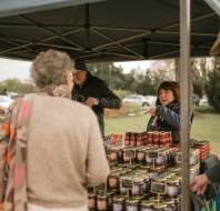 A woman stands behind a market stall selling jars of preserves to three customers outdoors under a canopy.