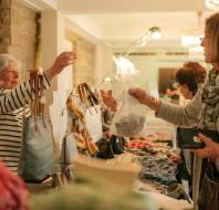 A woman behind a table sells bags and accessories to another woman at an indoor market.