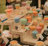 Colorful candles in decorative glasses and teacups are displayed on white wooden shelves at a market stall.