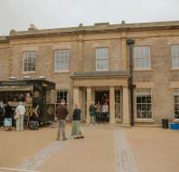 A food truck is parked outside a large stone building, with several people lined up to order; others stand or walk nearby, and a table with items is set up to the right.