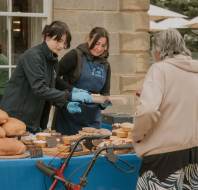 Two vendors behind a table of baked goods serve a customer using a walker at an outdoor market stall.