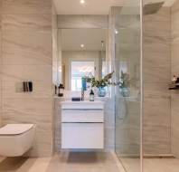Modern bathroom with beige tile walls, wall-mounted toilet, white vanity with mirror, and glass shower enclosure. Decorative plants on the vanity and a bottle on a shower shelf.