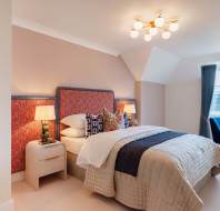 A bedroom featuring a neatly made bed with red patterned headboard, two bedside tables with lamps, a blue chair, a desk near a window, and a modern ceiling light.