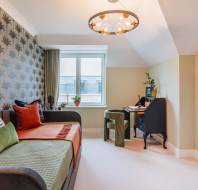 Room with a daybed, decorative pillows, a desk, chair, and lamp. Wallpaper with a leaf pattern on one wall, window with a view, and a mix of green and orange accents.