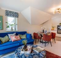 Living room with a blue sofa, colorful cushions, and a patterned tablecloth on the coffee table. A dining area with four red chairs is visible, and a kitchen is in the background.