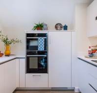Modern kitchen with white cabinets, a double oven, and minimalist decor. A wooden cutting board, potted plants, and decorative plates adorn the space.