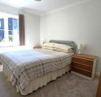 A bedroom with a double bed, two bedside tables with lamps, a wooden dresser, beige carpet, and a window with brown curtains letting in natural light.