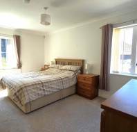 Bright bedroom with a double bed, wooden furniture, two windows with beige curtains, and neutral-colored walls and carpet.