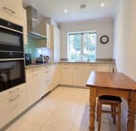 Modern kitchen with cream cabinets, built-in oven and microwave, wooden table with a stool, window, and a wall clock.