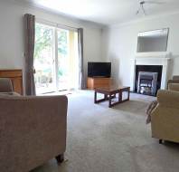 A living room with beige sofas, a coffee table, TV, fireplace, mirror, and sliding glass doors leading outside. Light carpet and neutral walls.