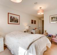 A neatly made bed in a bright bedroom with white walls, bedside tables, lamps, framed artwork, a pink armchair, and a teddy bear on a chest of drawers.