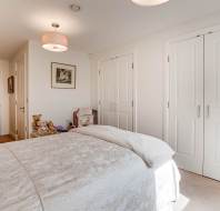 A neatly made bed in a bright bedroom with white walls, double closet doors, framed art, and stuffed animals on a chair.