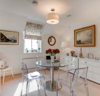 A bright, tidy room with a glass dining table, four clear chairs, white dressers, floral decor, lamps, wall art, and a window with a patterned roman shade.