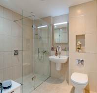Modern bathroom with beige tiles, glass-enclosed shower, wall-mounted sink, toilet, mirror, and shelves holding toiletries and a decorative flower.