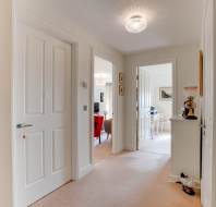 A hallway with beige carpet and white walls leads to multiple rooms, including a dining area with red chairs; a small cabinet and light fixture are visible.