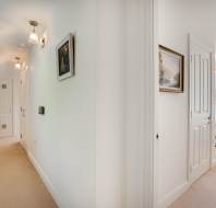 A hallway with beige carpet leads to a white door; another corridor leads to a bright bedroom with white bedding and floral window blinds.
