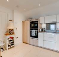 Modern kitchen with white cabinets, built-in oven, stove, kettle, countertop appliances, a wine rack, and a lamp on a small table. The room is well-lit with recessed ceiling lights.