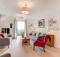 Bright living room with two grey sofas, a red armchair, coffee table, lamps, wall art, and large windows with floral curtains letting in natural light.
