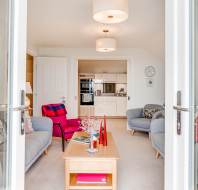 Bright living room with gray sofas, pink chairs, a wooden coffee table, and an open view into a modern white kitchen. Ceiling lights and floral curtains are visible.