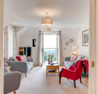 Bright living room with two grey sofas, a red armchair, coffee table, TV, and large window with floral curtains letting in natural light.