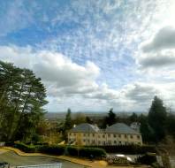 A view of buildings surrounded by trees under a partly cloudy sky, with a distant landscape visible on the horizon.