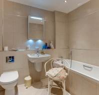 Modern bathroom with beige tiles, wall-mounted sink, toilet, bathtub, illuminated mirror cabinet, chair with cushions, and toiletries on the edge of the tub.