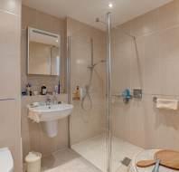 Modern bathroom with beige tiles, wall-mounted sink, glass shower enclosure, assorted toiletries on the sink, and a toilet with a bin and toilet paper nearby.