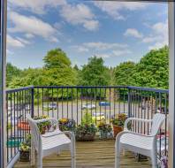 A small balcony with two white plastic chairs, several potted plants, and a view of a parking lot with cars and trees under a blue sky with clouds.