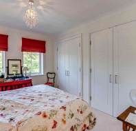 A bedroom with a floral bedspread, two windows with red blinds, a double-door closet, and a small desk with lamps and a mirror.