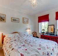 A neatly made bed with a floral duvet, two bedside tables with lamps, framed pictures on the wall, and a vanity table under two windows with red blinds.