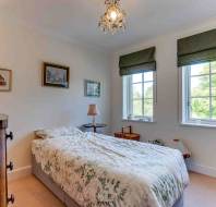 A neatly made single bed in a bright bedroom with two windows, green roman shades, framed wall art, a chest of drawers, and a bookshelf.