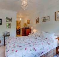 A neatly made bed with a floral duvet, surrounded by side tables with lamps, framed pictures on white walls, and a wooden dresser in a well-lit bedroom.