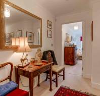 A tidy hallway with a wooden dressing table, two chairs, a lamp, framed mirror, and various decorative items; a bedroom is visible through an open door.