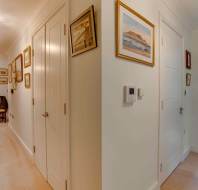 A well-lit hallway with cream walls and carpet, decorated with framed pictures, leads to a bedroom with a window and green valance. Closed doors are visible along the corridor.