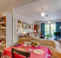 A cozy living room with patterned sofas, a red dining table, bookshelves, framed artworks on the walls, and large windows with blue curtains letting in natural light.