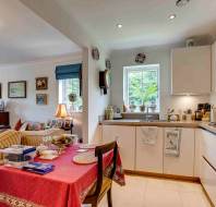 Bright, tidy kitchen and living area with white cabinets, a window over the sink, a table set for a meal, and cozy seating with framed art on the walls.
