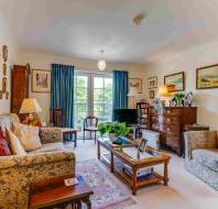 A traditionally furnished living room with two patterned sofas, a coffee table, framed art on the walls, a chest of drawers, and a window with blue curtains letting in natural light.