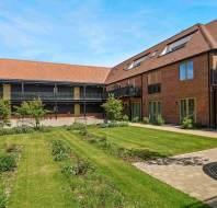 A modern brick building with balconies faces a landscaped courtyard with green lawns, young trees, and a circular wooden bench under a blue sky.