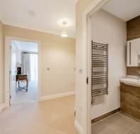A view of a hallway with beige carpet leading to a bright room and an open bathroom featuring a sink, mirror, heated towel rail, and tiled wall.