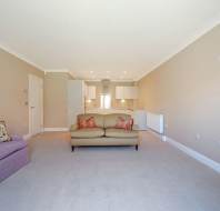 A spacious living room with beige walls, light carpet, a lavender sofa, a beige loveseat, and an open-plan kitchen in the background.