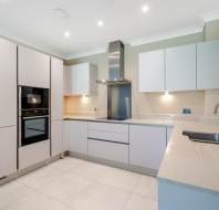 Modern kitchen with light gray cabinets, built-in oven, stainless steel range hood, under-cabinet lighting, and a single-basin sink on a beige countertop.