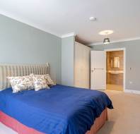 A bedroom with a double bed covered in a blue duvet, floral pillows, light green walls, a white wardrobe, and an open door leading to a bathroom.
