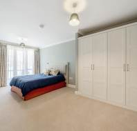 A bedroom with a double bed, large white wardrobe, beige carpet, light green walls, and a window with curtains letting in natural light.